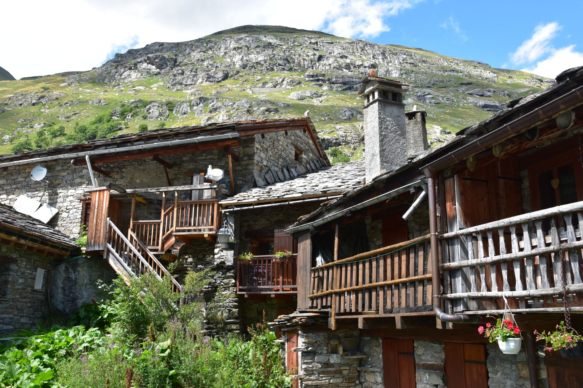 Maisons de village en pierre et bois avec balcons fleuris, devant une montagne rocheuse et herbeuse sous un ciel bleu.