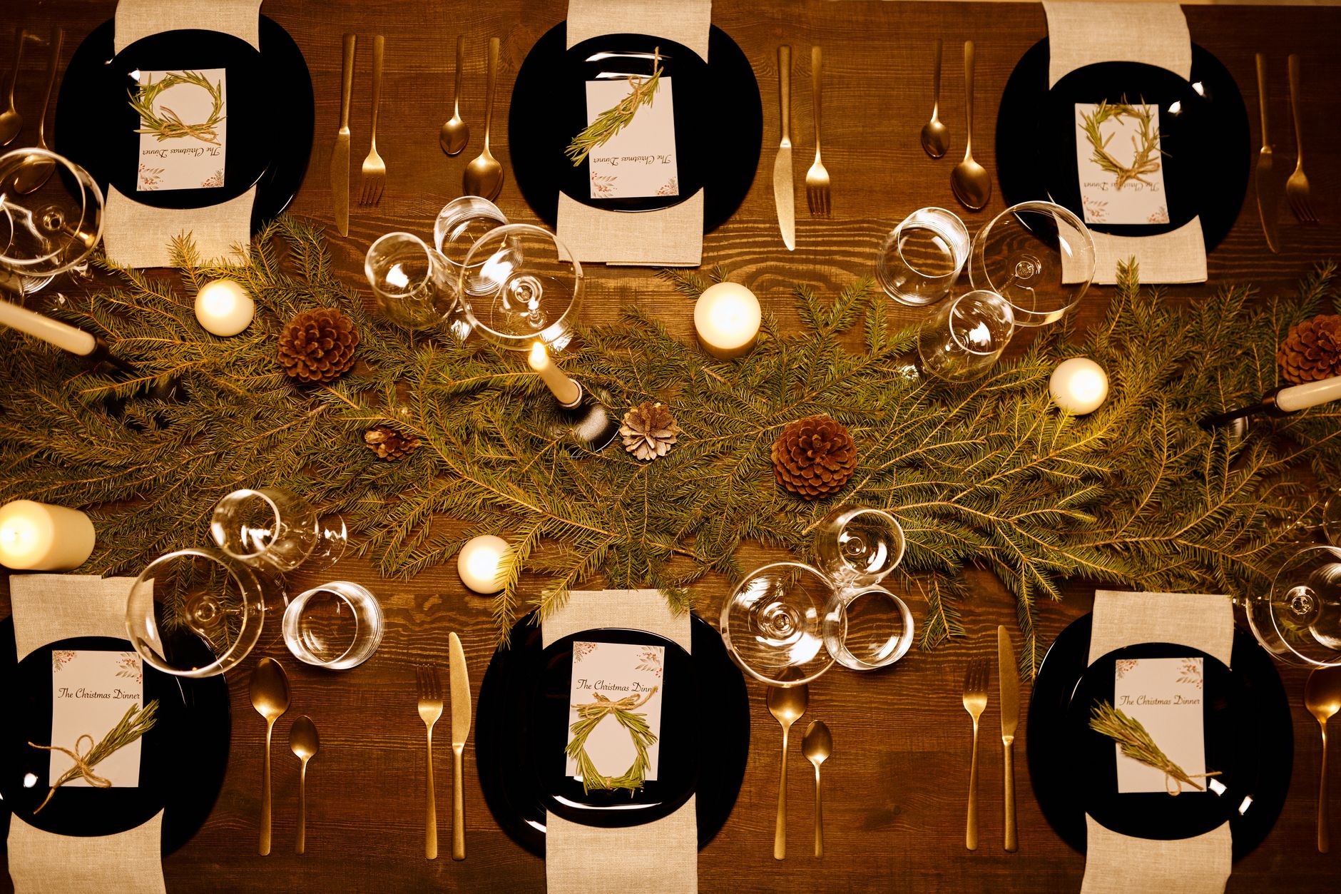 Table de fête vue du dessus : assiettes noires, serviettes en lin, couverts dorés, verres, bougies et branches de sapin.