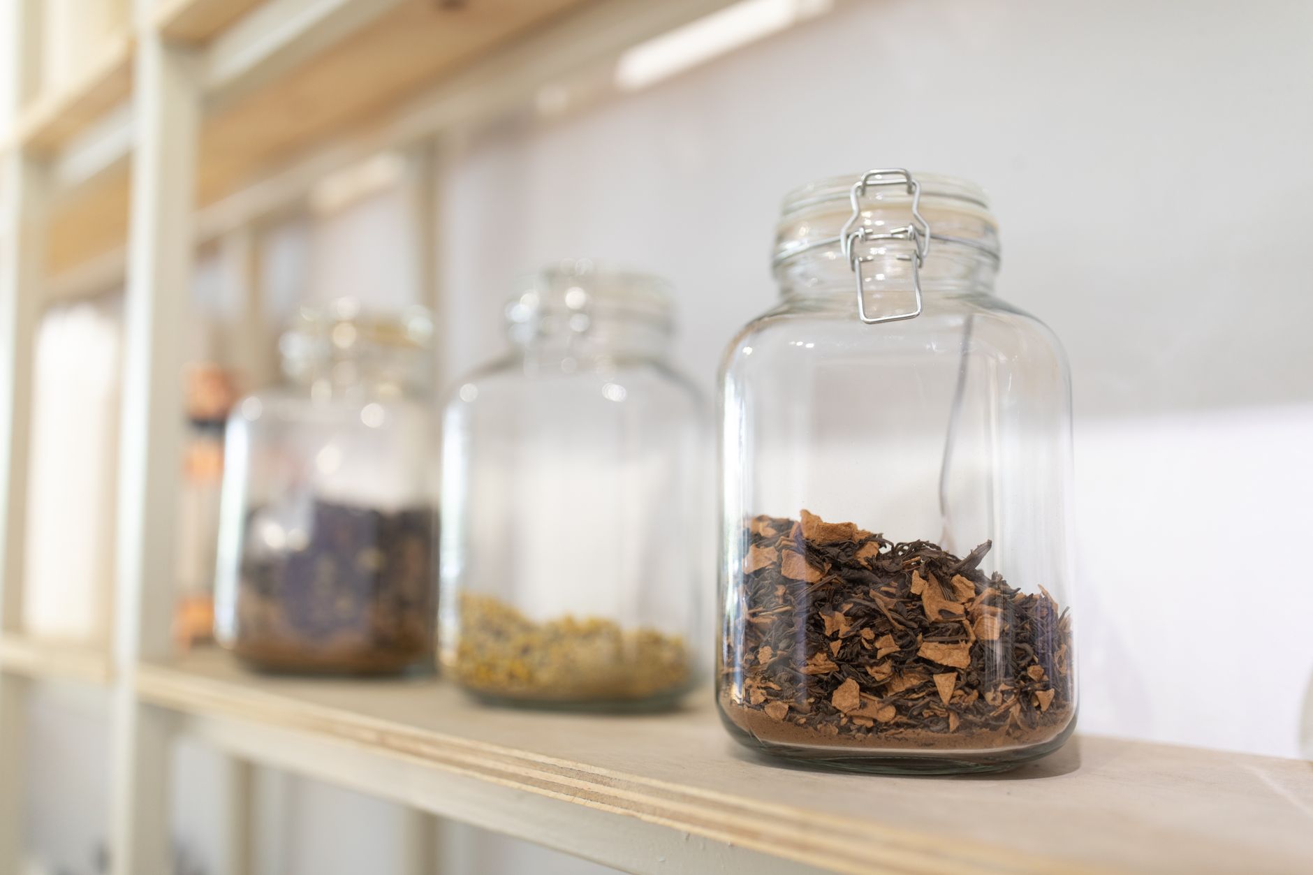 Trois bocaux en verre à fermeture métallique sur une étagère en bois clair, celui du premier plan contenant des feuilles séchées brunes.